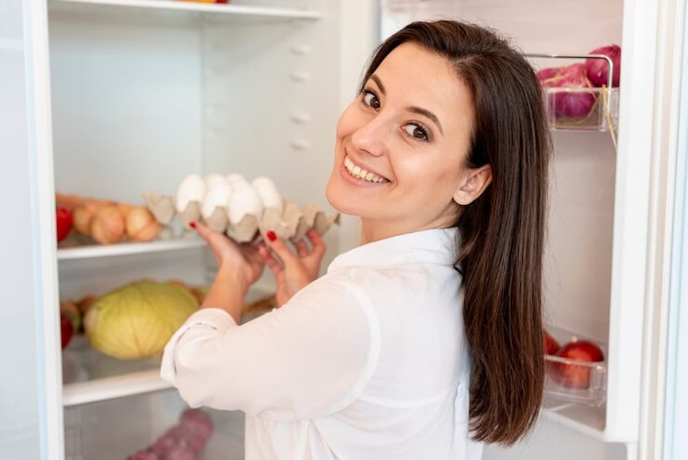 Mujer sonriente sosteniendo cartón de huevos junto al frigorífico